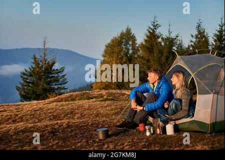 Vue latérale de couple de voyageurs mariés se reposant dans une tente sur un pré de montagne pendant la randonnée d'automne sur la toile de fond des arbres et de la colline de montagne. Matin de soleil dans le camping. Banque D'Images