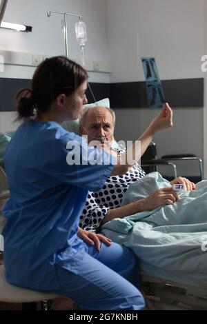 Patient âgé souffrant d'une maladie des poumons qui respire en utilisant un masque à oxygène posé dans le lit d'hôpital, infirmier écoutant montrant un diagnostic d'expose de rayons X avant la chirurgie. Banque D'Images
