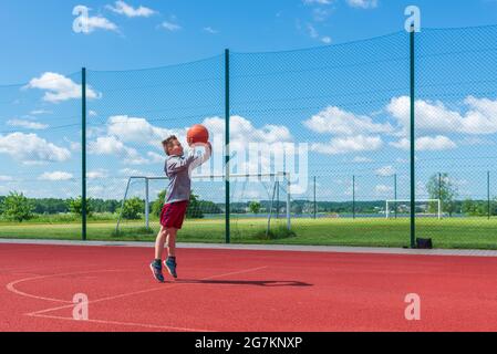 Joli petit garçon de tir balle au cerceau à l'aire de jeux.mignon jeune garçon joue au basket-ball sur l'aire de jeux en été chaud jour. Banque D'Images