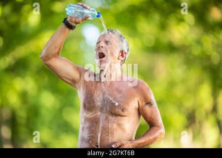 Un vieil homme sans chemise verse de l'eau dans une bouteille au-dessus de sa tête et de son visage à l'extérieur lors d'une chaude journée d'été. Banque D'Images