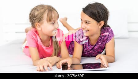 Deux petites filles mignonnes utilisent une tablette numérique et sourient lorsqu'elles sont allongées sur le lit dans la chambre des enfants Banque D'Images