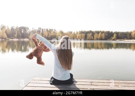 Une fille heureuse joue avec un ours en peluche assis sur la jetée près de la rivière Banque D'Images