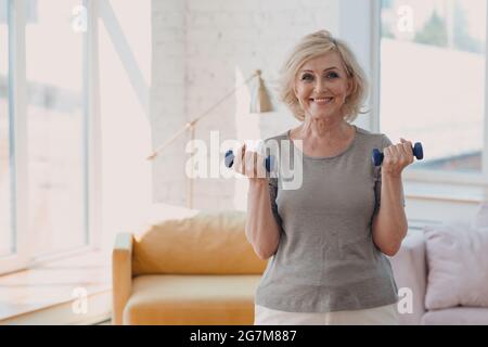 Vieux caucasien vieille femme âgée portrait gris cheveux faire des exercices avec des haltères dans l'habillement décontracté à la maison appartement salon Banque D'Images