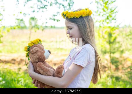 Une fille avec une couronne de pissenlits regarde l'ours en peluche dans le parc Banque D'Images