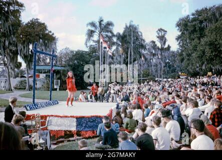 Une adolescente, tout vêtue de rouge, se produisant sur une scène extérieure sous le soleil de la soirée à un concours ‘miss gailler USA’ à Cypress Gardens, dans le comté de Polk, en Floride, aux États-Unis en 1968. Elle chante (ou applaudit ou chante) accompagnée d'un groupe (en bas à gauche), avec des photographes présents devant un public important. Cette image provient d'une ancienne transparence couleur amateur Kodak américaine, une photographie vintage des années 1960. Banque D'Images