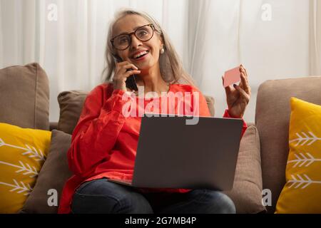 Une vieille femme parle au téléphone avec une carte à la main et un ordinateur portable sur les genoux. Banque D'Images