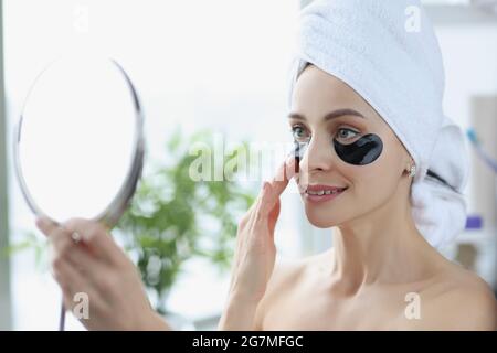 Jeune femme avec des taches noires sous les yeux et avec une serviette sur sa tête regardant dans le miroir à la maison Banque D'Images