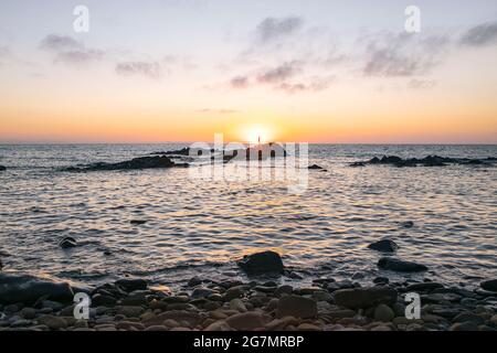 Coucher de soleil sur la plage italienne, côte rocheuse. Un voilier traverse le disque solaire. Réflexions du soleil sur la surface de l'eau. Banque D'Images