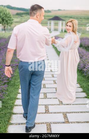 Donnez-moi votre main, suivez-moi. L'amour à travers les années ensemble. Jolie charmante blonde mature dame dans une élégante robe, tenant la main et marchant avec son beau homme dehors dans le champ de lavande avec belvédère Banque D'Images