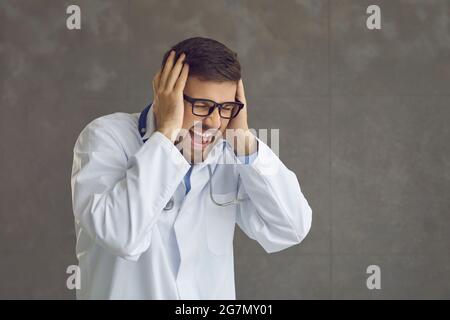 Un médecin stressé en colère crie et ferme les oreilles avec les mains isolées sur fond gris Banque D'Images