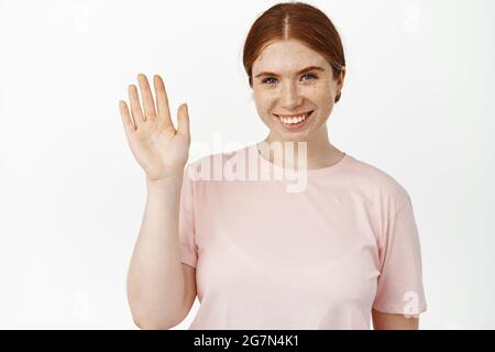 Gros plan portrait d'une fille au gingembre sympathique avec un sourire sincère, en agitant la main à l'appareil photo, dites bonjour, saluant les clients, vous accueillir, faites un geste de vague Banque D'Images