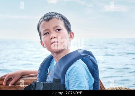 Un garçon asiatique sérieux en gilet de sauvetage sur bateau de plaisance sur la mer. Banque D'Images