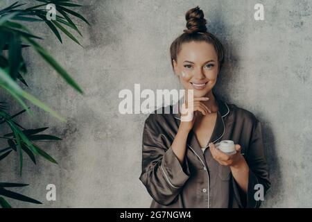 Happy female porter un pyjama brun en soie avec crème hydratante dans sa main pendant la routine de beauté du matin Banque D'Images