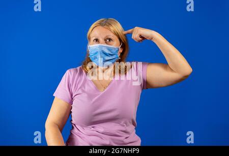 Jolie femme d'âge moyen dans un t-shirt violet et un masque chirurgical pour se protéger contre le coronavirus avec ses index sur sa tête isolés en bleu Banque D'Images