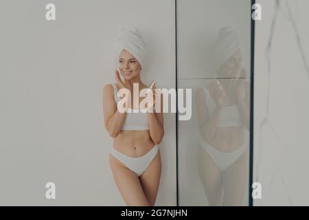 Belle femme debout dans la salle de bains après la douche du matin et l'application de crème hydratante sur le visage Banque D'Images