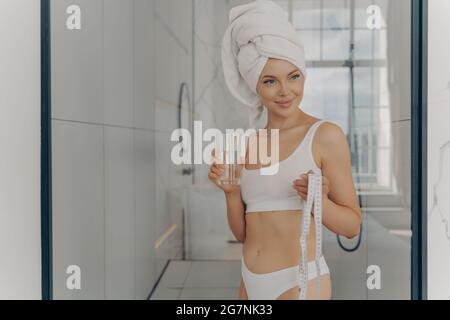 Jeune fille sportive attrayante tenant un ruban de mesure et se rafraîchissant avec un verre d'eau minérale dans la salle de bains Banque D'Images