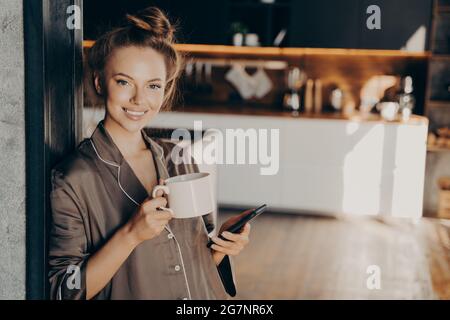 Portrait d'une jeune femme positive tenant un smartphone à la main et buvant du café tout en étant debout dans la chambre à coucher Banque D'Images