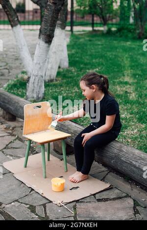 jolie fille peint une chaise en bois avec de la peinture jaune, un passe-temps écologique pour les enfants Banque D'Images