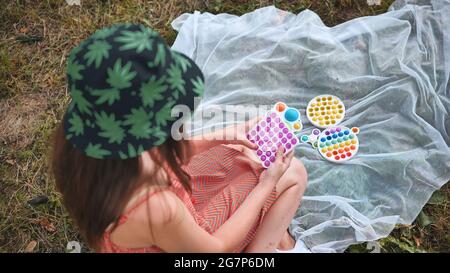 Une jeune fille joue avec un popit anti-stress et des jouets simples à fossettes dans le parc lors d'une journée d'été. Banque D'Images