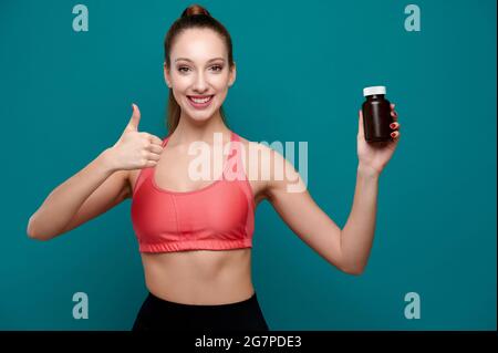 Souriant jeune caucasienne entraîneur de forme physique femelle tient des pilules dans une bouteille et montre pouce vers le haut Banque D'Images