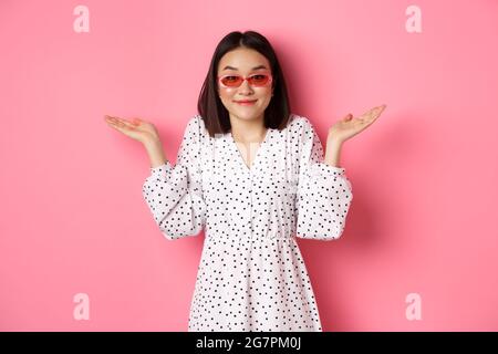 Mignonne asiatique touriste souriant à la caméra, haussant l'indice, ne sais pas, portant des lunettes de soleil à la mode et robe blanche, debout contre fond rose Banque D'Images