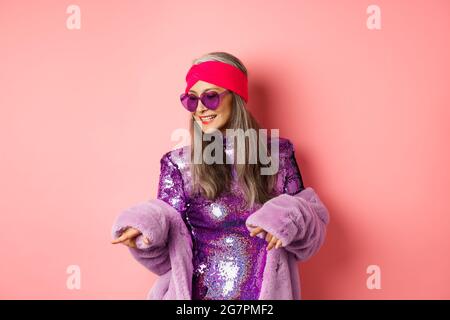 Femme asiatique à la mode dansant disco, portant une robe brillante étincelante et des lunettes de soleil en forme de coeur, s'amusant, debout sur fond rose Banque D'Images
