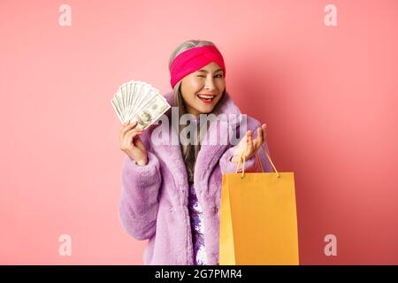 Femme asiatique riche et à la mode gaspillant de l'argent dans les magasins, tenant sac de shopping et dollars, en se faisant un plaisir à l'appareil photo, fond rose Banque D'Images