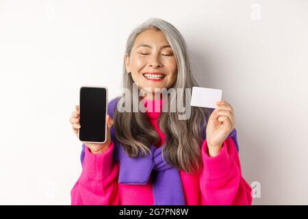 Achats en ligne. Heureuse et ravie asiatique femme senior souriant avec joie, montrant l'écran vide de smartphone et carte en plastique, l'achat dans Internet, blanc Banque D'Images