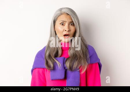 Gros plan d'une femme asiatique âgée choquée qui resplense, regardant avec un visage confus, regardant avec des cringe, se tenant sur un fond blanc Banque D'Images