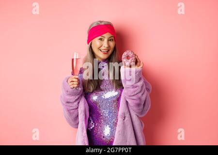 Belle et élégante femme asiatique senior s'amuser, boire du champagne et manger du beignet sucré, sourire à l'appareil photo, debout sur fond rose Banque D'Images