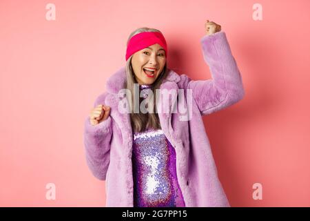 Concept commercial. Joyeuse femme asiatique dansant et s'amusant, portant un élégant manteau et une robe en fausse fourrure pourpre, gagnant le prix et souriant Banque D'Images