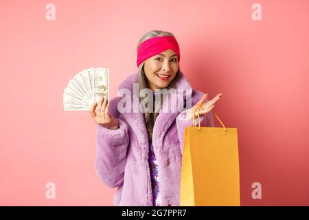 Une grand-mère riche et élégante qui fait du shopping avec des dollars, tient un sac en papier et sourit sans souci, se tenant sur un fond rose Banque D'Images