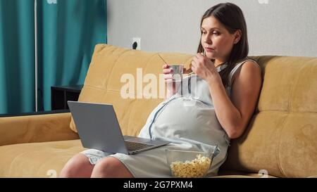 Femme enceinte qui mange du yaourt et du pop-corn et qui regarde un ordinateur portable tout en étant assise sur un canapé. Banque D'Images