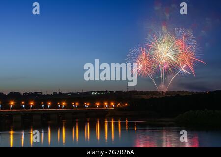 Feux d'artifice sur le fond d'un ciel clair de nuit près de la rivière dans laquelle la lumière est réfléchie et le pont à travers cette rivière Banque D'Images