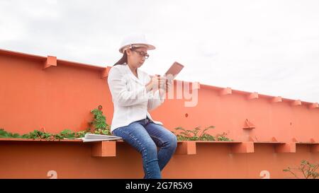 Femme ingénieur dans un casque blanc parlant radio de communication (talkie walkie) et tenant une tablette numérique, se tient dans le chantier de construction de l'hydroel Banque D'Images