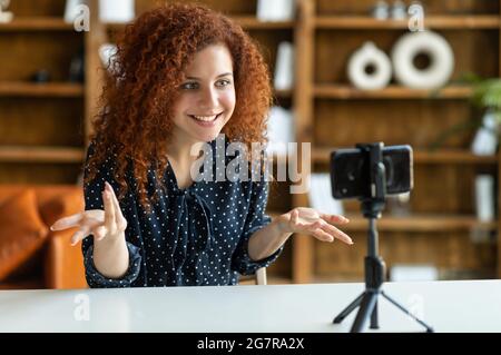 Une femme curieusement attirante parlant à l'appareil photo sur le téléphone sur un trépied, en faisant des gestes, en blogging ou en parlant en direct, en enregistrant une présentation vidéo, en tenant un appel vidéo sur le téléphone mobile Banque D'Images