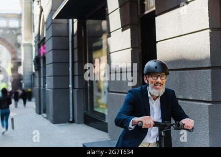 joyeux homme d'affaires mature dans blazer et casque à cheval e-scooter Banque D'Images