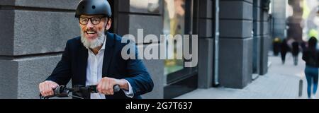 joyeux homme d'affaires mature dans blazer et casque à cheval e-scooter, bannière Banque D'Images