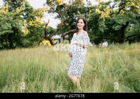 Jolie femme caucasienne en robe d'été caressant son ventre enceinte en posant au jardin vert. Concept d'attente et grossesse en santé. Banque D'Images