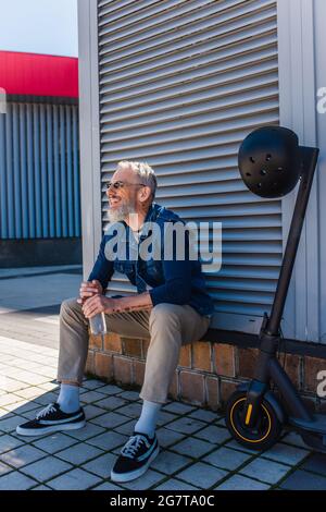 homme mature barbu en lunettes de soleil souriant et tenant une bouteille d'eau près d'un e-scooter avec un casque Banque D'Images