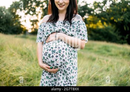 Gros plan de la femme blanche enceinte en robe d'été posant à l'extérieur. Jolie brune caressant son ventre tout en se tenant dans un jardin verdoyant. Banque D'Images