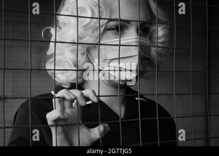 triste femme blonde peur dans le masque derrière les barres gros plan portrait, monochrome Banque D'Images