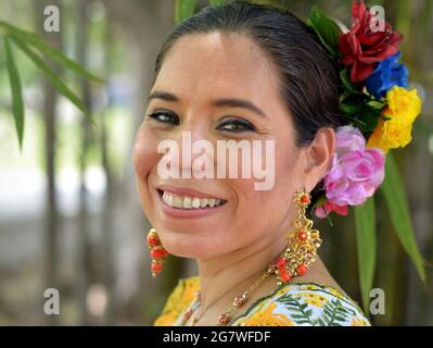 La jeune femme yucatecan mexicaine porte une robe folklorique maya traditionnelle avec de longues boucles d'oreilles et des fleurs colorées dans ses cheveux à l'arrière-plan du parc. Banque D'Images