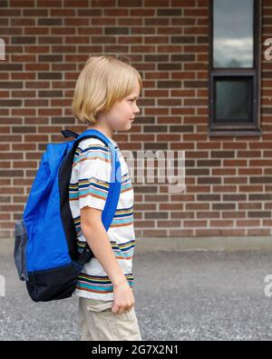 Beau enfant près du mur de briques de bâtiment d'école à la cour d'école. Petit élève heureux portant un sac à dos bleu, marchant jusqu'à l'école. Banque D'Images