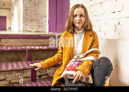 Portrait d'une jeune adolescente assise sur un escalier violet contre un mur de briques blanches. Banque D'Images