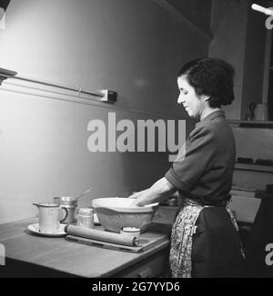 années 1950, historique, une dame dans une robe avec un tablier, debout sur une table en bois dans un coin d'une pièce mélangeant de la pâte dans un bol de farine, sur un plateau de pâtisserie en bois. Avec son rouleau à côté d'elle et quand elle est prête à la pâte, elle est prête à faire de la pâtisserie, Angleterre, Royaume-Uni. Banque D'Images