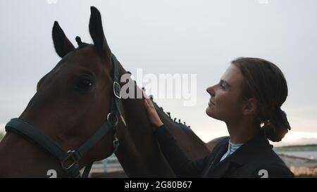 Une horsewoman qui lui sourit en lui caressant son cheval brun. La fille est vêtue d'un manteau noir. Regarde son cheval avec amour. Ciel blanc clair en arrière-plan. Amour pour les chevaux. Banque D'Images