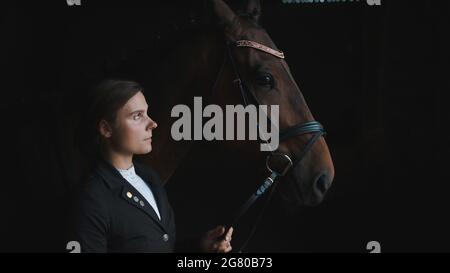 Horsewoman posant avec son cheval brun joint tenant son bride. La fille est vêtue d'un manteau noir. Le cheval porte des bijoux de tête. Toilettage de chevaux. Liaison entre cheval et son maître. Banque D'Images