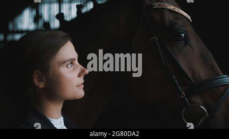 Horsewoman posant avec son cheval brun de phoque. La fille est vêtue d'un manteau noir. Le cheval porte des bijoux de tête. Toilettage de chevaux. Liaison entre cheval et son maître. Banque D'Images