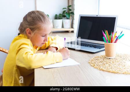 Une écolière caucasienne fait ses devoirs à la maison, écrit dans un carnet. Fille de 10-11 ans avec ordinateur portable. Enseignement à domicile, e-learning. Retour à l'école. Confortable Banque D'Images
